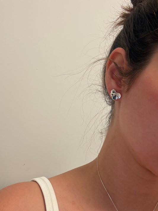 A close-up side profile of a person wearing a small clear faceted stud earring on their ear, with dark hair pulled back and a white strap visible at the shoulder against a plain light background.