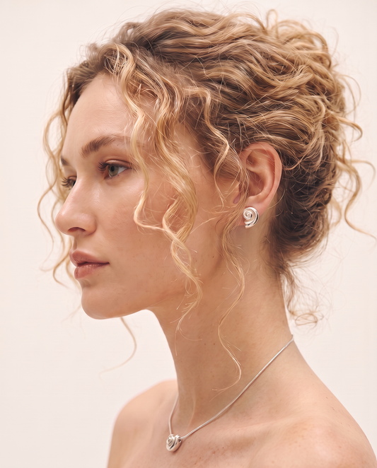 A woman in profile wearing a silver spiral-shaped stud earring and matching silver necklace against a plain light background.