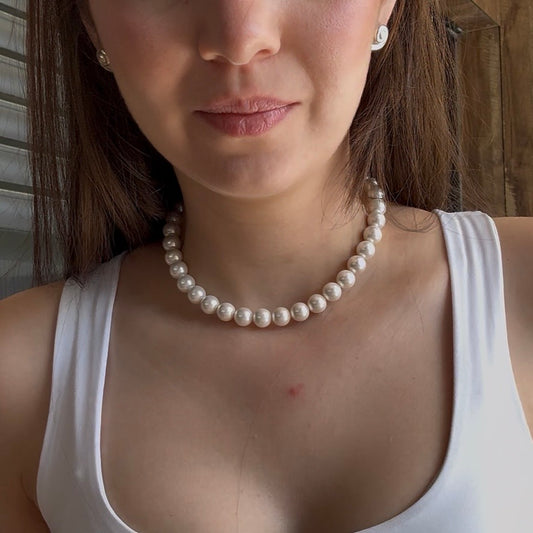 A close-up of a woman wearing a classic white freshwater pearl necklace with matching pearl stud earrings, styled with a white tank top.